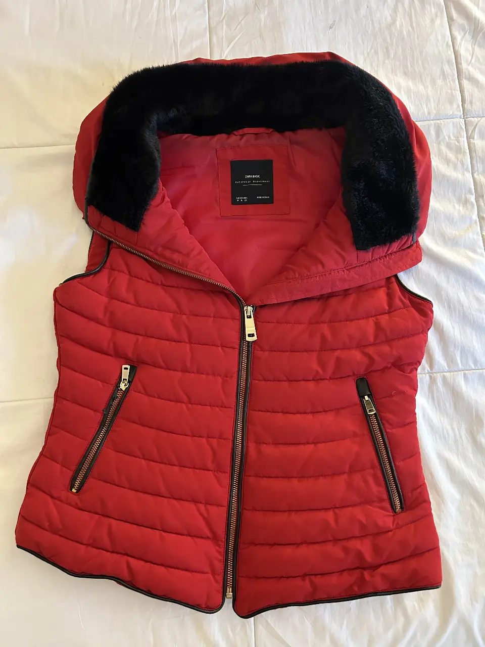 Chaleco Zara acolchado rojo con capucha y borde de piel sintética negra. Cierre con cremallera frontal y bolsillos laterales con cremallera. Ideal para el invierno.