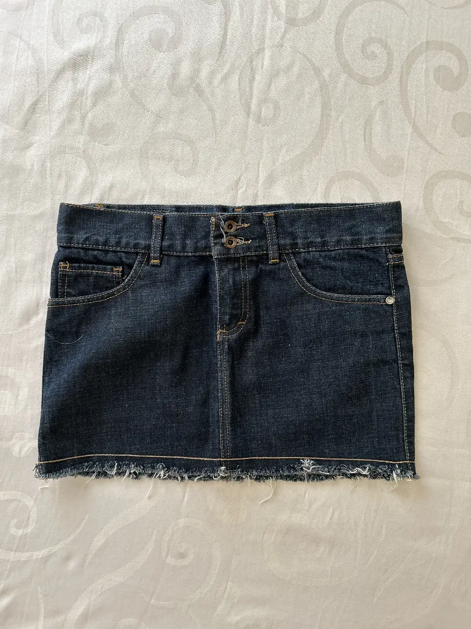 Minifalda de jean azul oscuro con detalles de desgaste en el borde inferior. Diseño clásico de cinco bolsillos y cierre con doble botón. Ideal para un look casual y moderno. Kosiuko.
