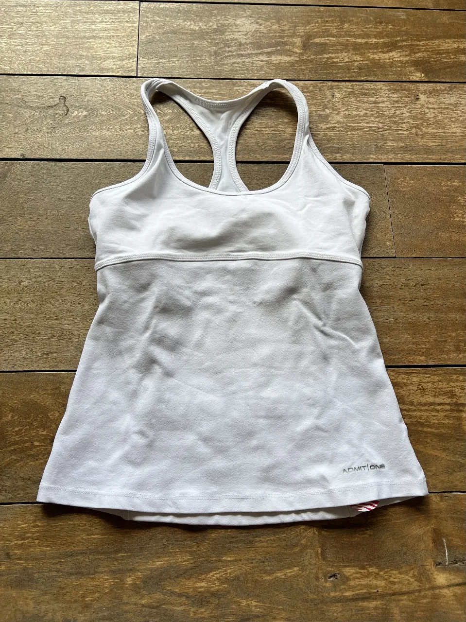 Top deportivo blanco de la marca Admit One, con diseño de espalda estilo nadador y detalle de bolsillo en la parte trasera. Ideal para actividades de running y gimnasia.