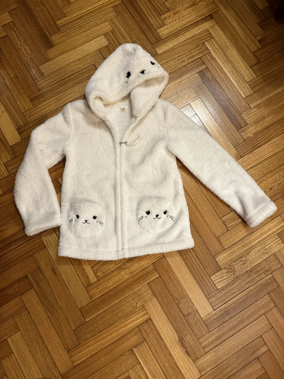 Buzo con capucha de peluche blanco con diseño de foca. Tiene cierre frontal y bolsillos delanteros con detalles de caritas de oso. Ideal para un look cómodo y divertido. y para arriba del pajama. 