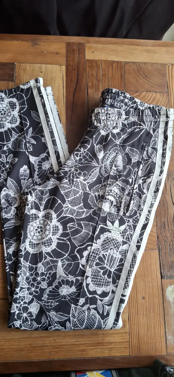Pantalón deportivo Adidas con estampado floral en blanco y negro. Diseño con las icónicas tres tiras blancas a los costados y cierre. Ideal para un look casual y cómodo.