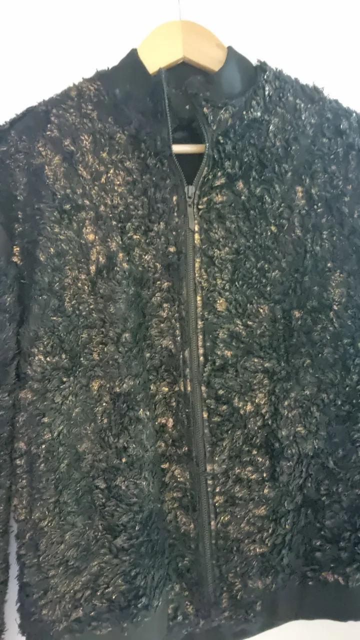 Campera bomber de mujer, negra con textura de pelo y detalles dorados. Cierre frontal y cuello redondo. Ideal para un look moderno y abrigado.