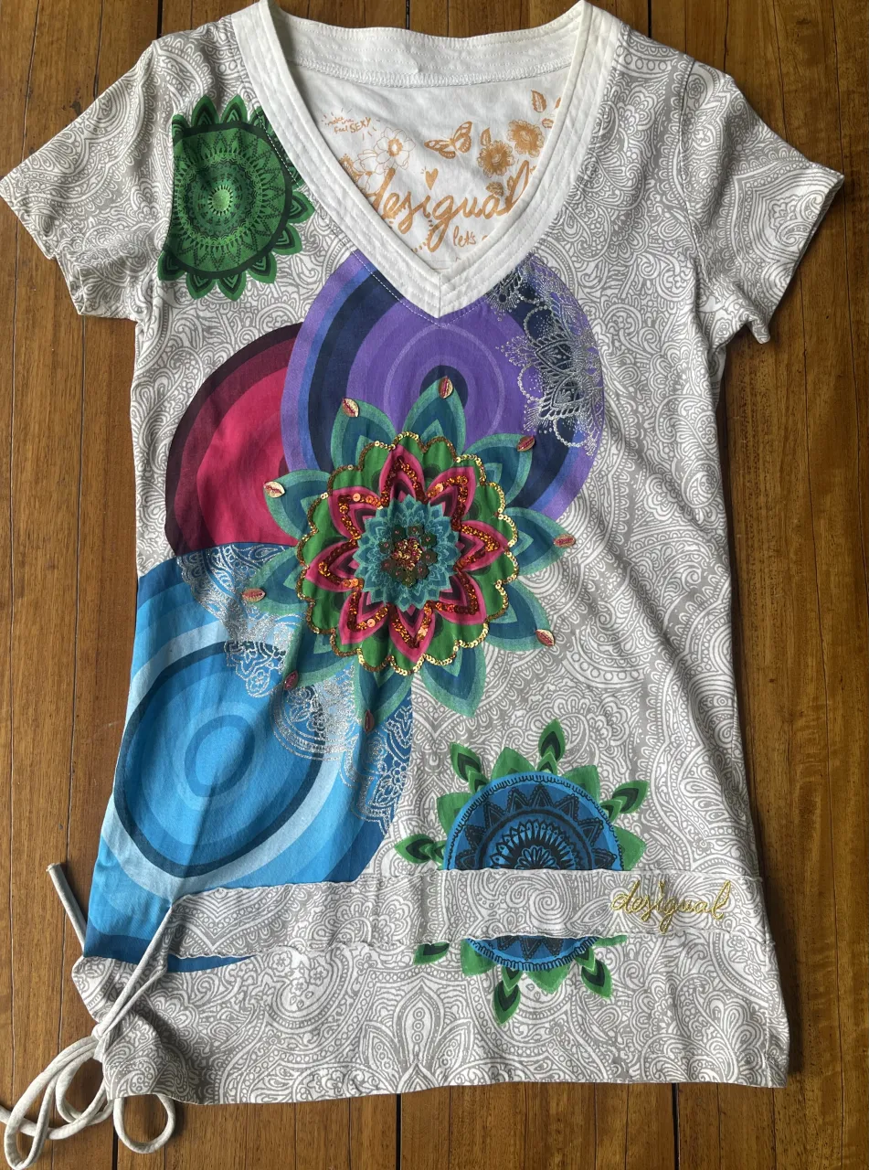 Remera manga corta con estampa floral y detalles de lentejuelas. Posee un diseño vibrante y colorido con un fondo gris con estampado paisley. Tiene un cordón ajustable en uno de los laterales para un calce personalizado. Ideal para un look casual y original.