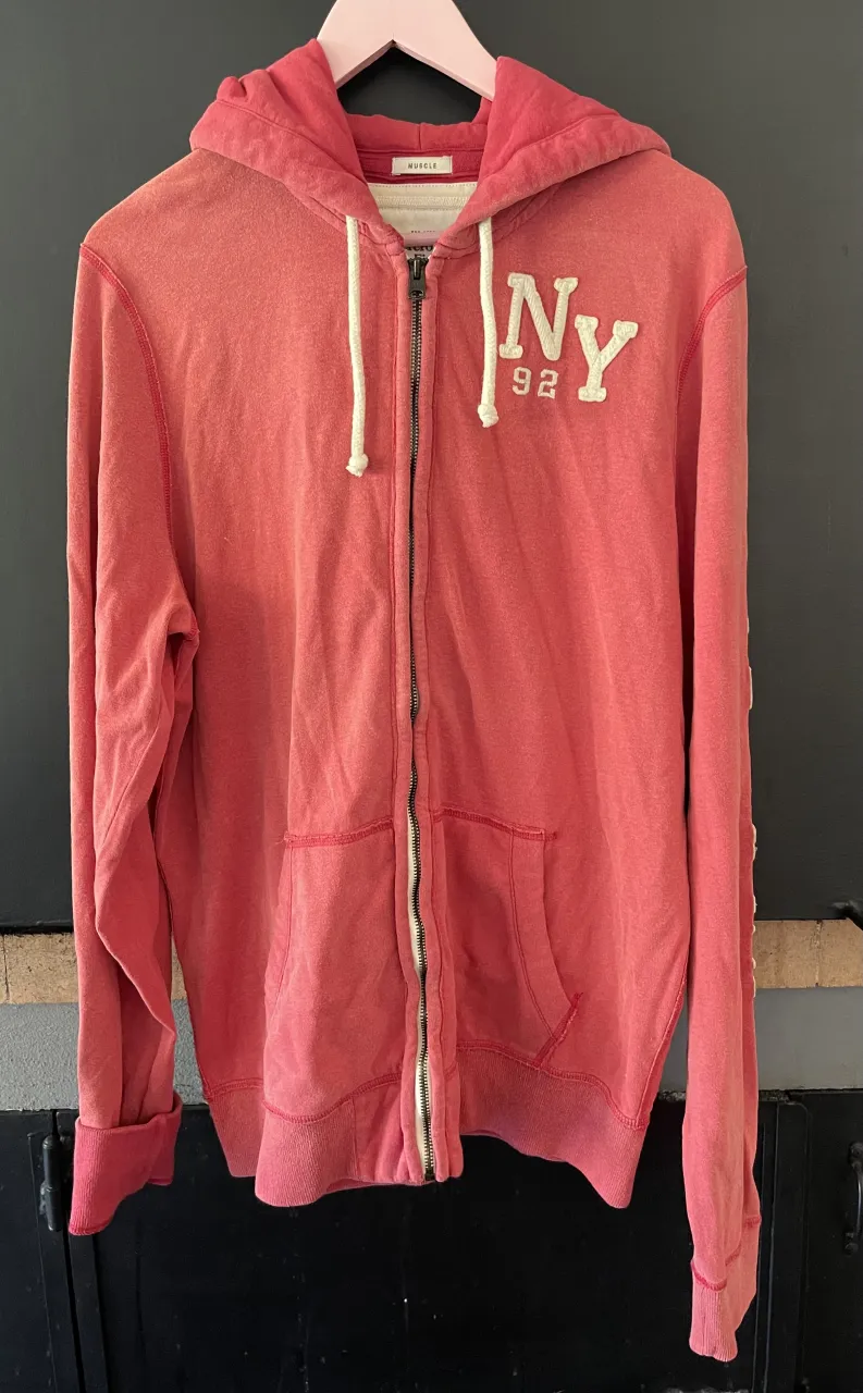 Buzo con capucha y cierre completo, color coral desgastado, con estampa "NY 92" en el frente y "Abercrombie & Fitch" en la manga. Ideal para un look casual y relajado.