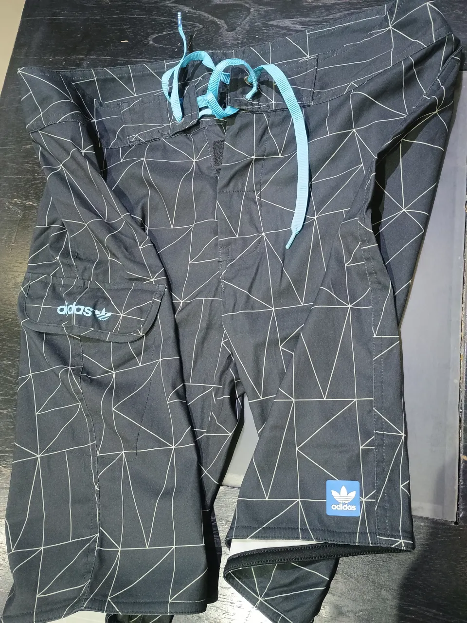 Short de baño Adidas negro con estampado geométrico y cordones celestes. Ideal para la playa o pileta, con bolsillo lateral y logo de la marca.