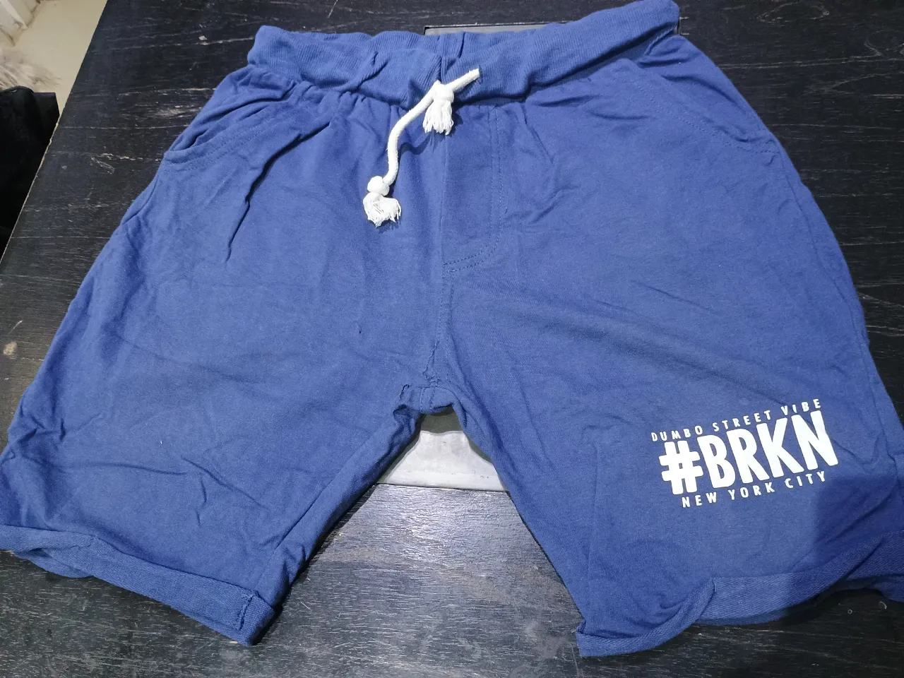Short de hombre de tela de algodón, color azul. Con cintura elastizada y cordón para ajustar. Tiene bolsillos laterales y un estampado en blanco con la frase "DUMBO STREET VIBE #BRKN NEW YORK CITY". Ideal para un look casual y cómodo.