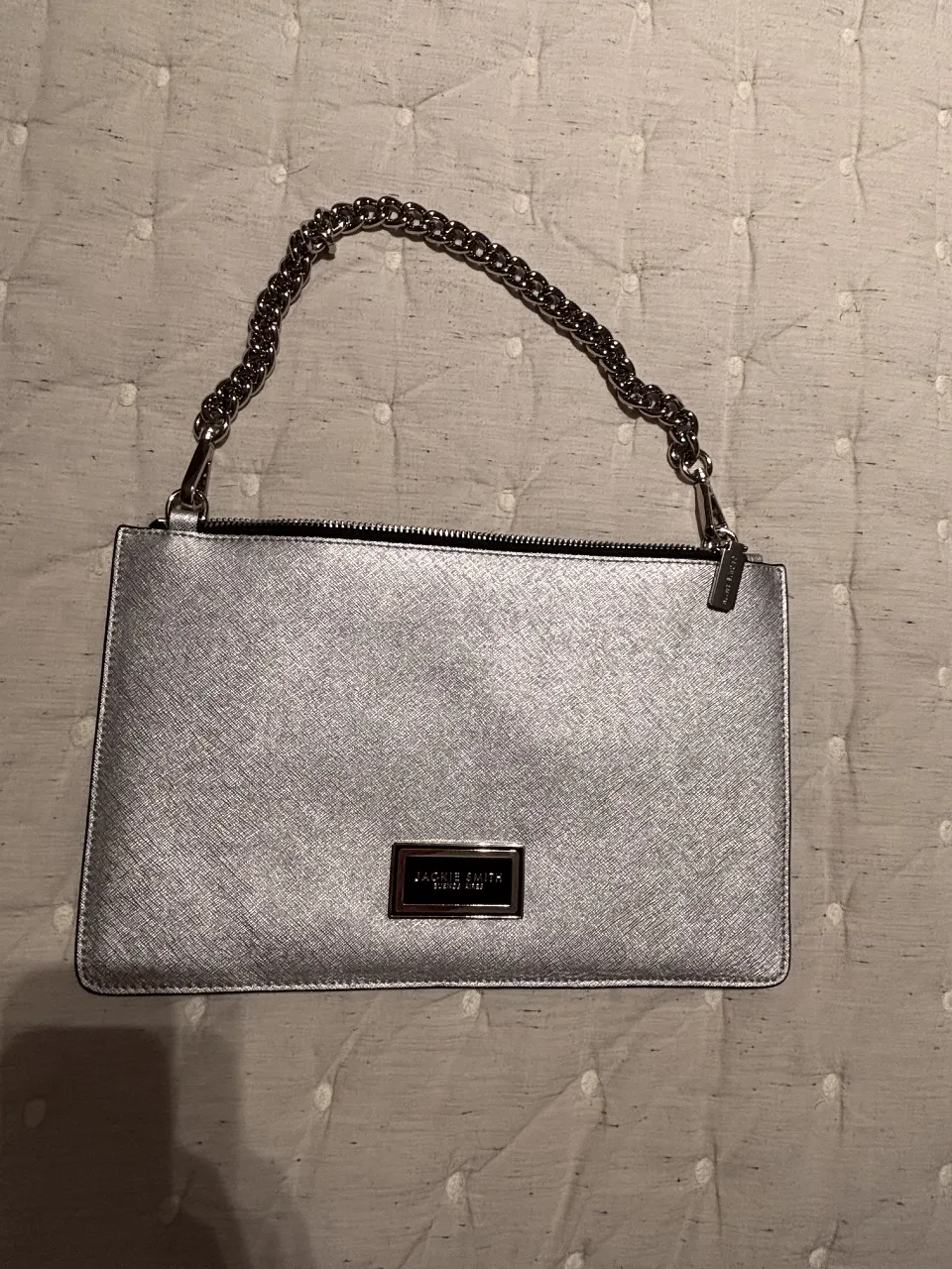 Cartera plateada de mano con cadena plateada. Tiene un aplique rectangular con el nombre de la marca. Ideal para una salida.