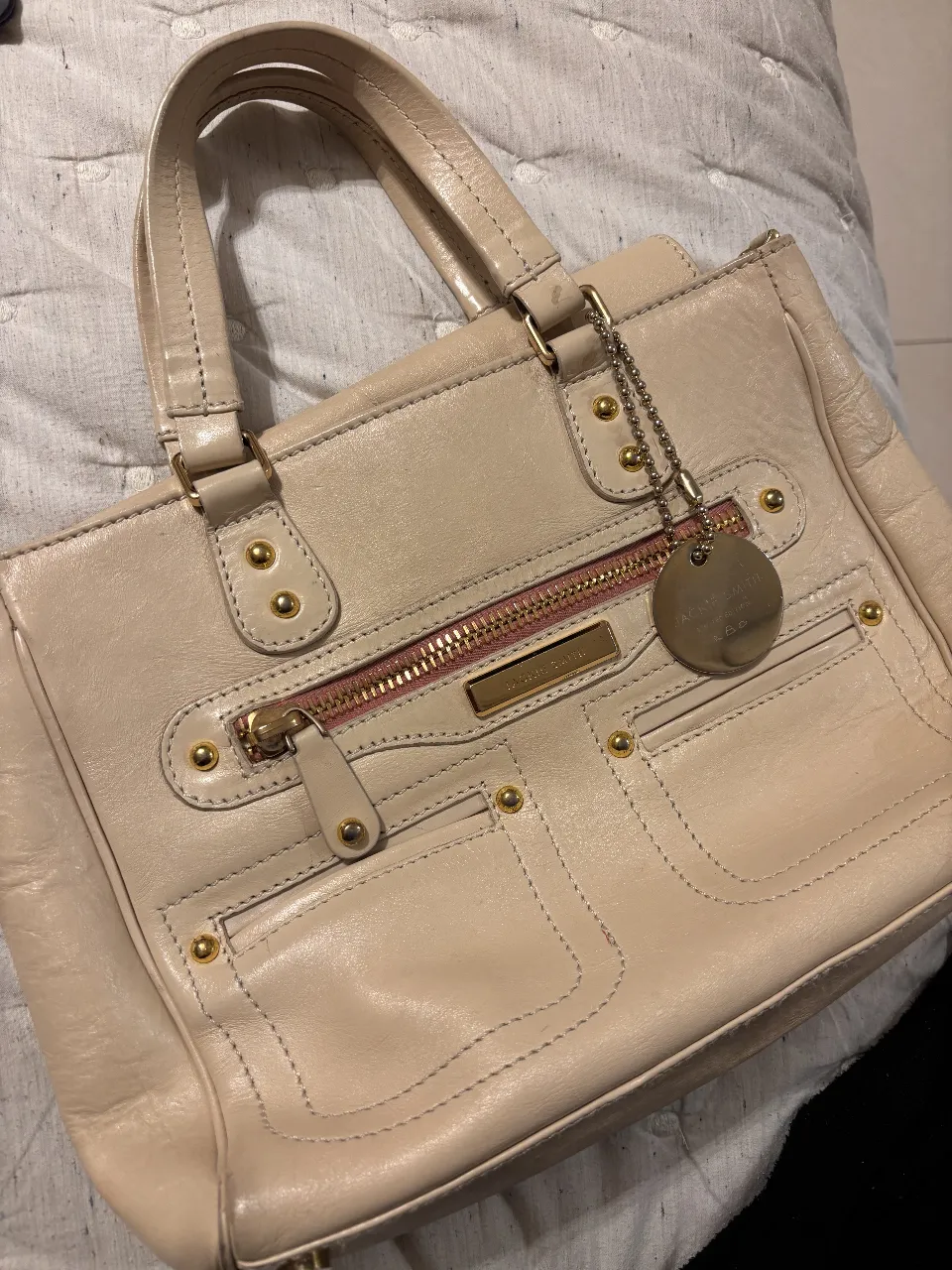 Cartera Jackie Smith de cuero en color crema, con detalles dorados y cierre con cremallera. Cuenta con dos manijas cortas y una correa larga desmontable. Interior forrado con tela a rayas.