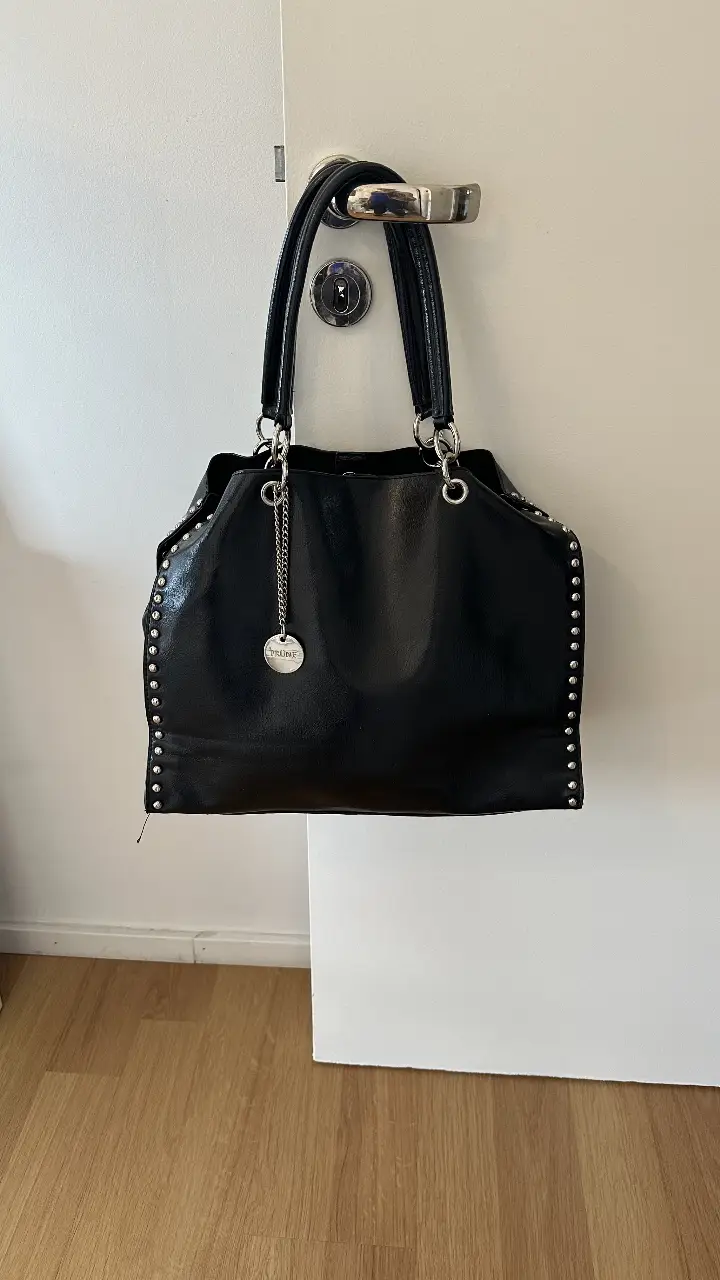 Cartera negra de cuero con tachas plateadas, marca Prüne. Diseño elegante con detalles metálicos y cadena colgante. Ideal para un look sofisticado.