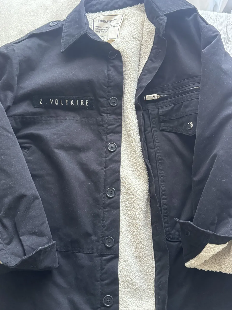 Campera negra con interior de corderito blanco. Tiene cuello de camisa, botones al frente y un bolsillo superior con cierre.