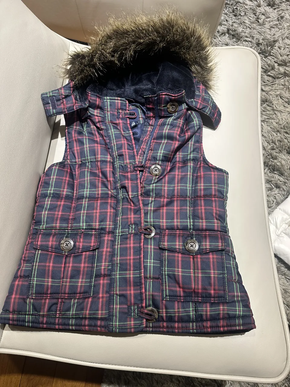 Chaleco acolchado para niña, con estampado a cuadros en tonos azul y rojo. Posee capucha con borde de piel sintética y cierre frontal con cremallera y botones. Ideal para el invierno.