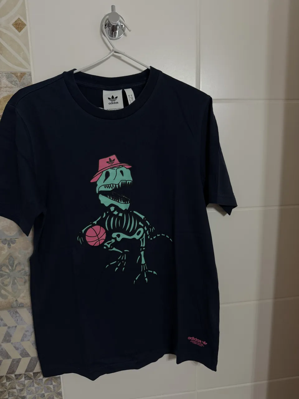 Remera de algodón azul marino con estampa de dinosaurio esqueleto con sombrero rosa y pelota de básquet. Logo de Adidas Originals en la parte inferior derecha y en la espalda.