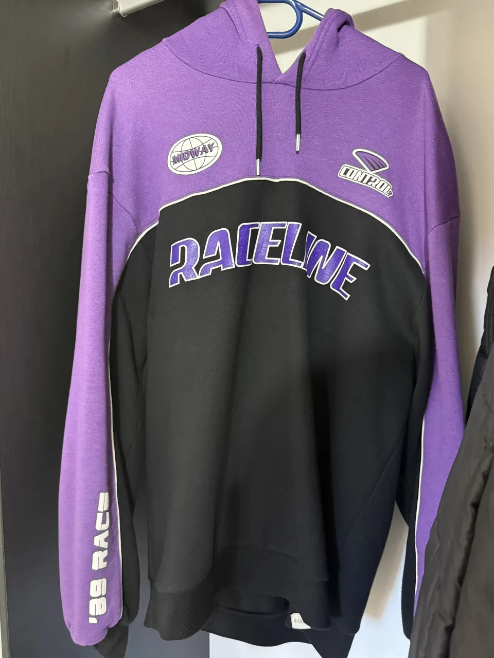Buzo con capucha de color púrpura y negro, con inscripciones "RACELINE" en el frente y "'89 RACE" en la manga. Posee detalles de ribetes blancos.
