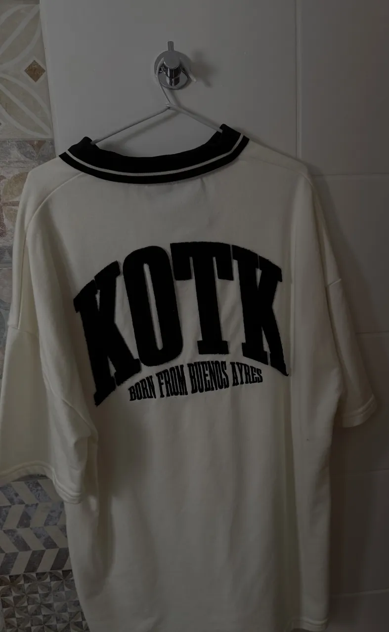 Remera blanca tipo polo con cuello y puños a rayas negras y blancas. En el frente tiene un bordado pequeño con la inscripción "kotk". En la espalda, tiene un bordado grande con "KOTK" y debajo "BORN FROM BUENOS AIRES". es bastante oversized casi un L te diría , tiene 2 usos 