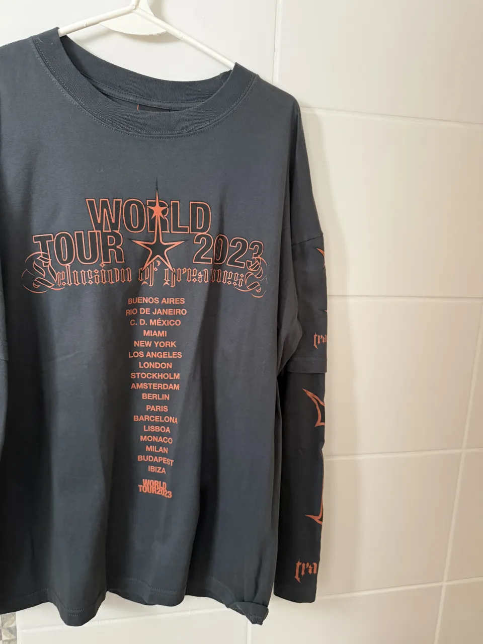 Remera manga larga gris oscuro con estampa "WORLD TOUR 2023" en naranja en el frente. La estampa incluye el nombre de varias ciudades. En las mangas tiene un diseño de "I" con la palabra "trapstar" debajo. En la espalda tiene un gran logo estampado.