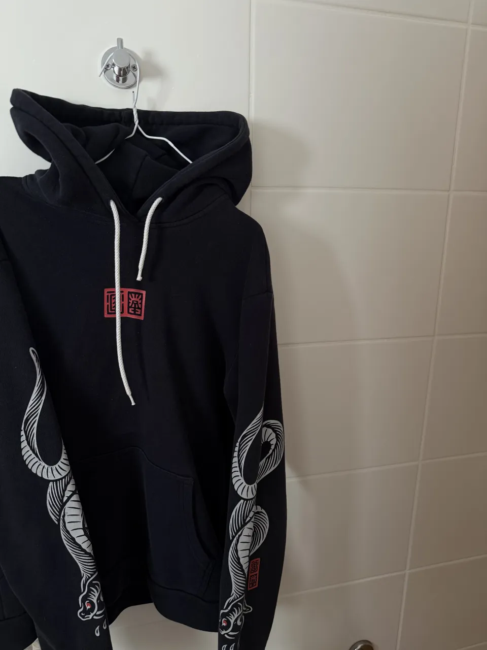 Buzo negro con capucha y cordones blancos. Posee un estampado de serpiente en las mangas y un logo cuadrado con caracteres rojos en el frente.