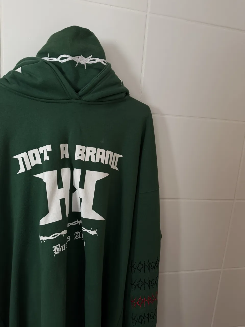 Buzo verde con capucha. Estampado blanco en la espalda con un círculo y el número 13 en el centro, rodeado de alambre de púas. En la manga izquierda tiene un estampado de rayos y en la manga derecha la palabra "KONGO" repetida varias veces, algunas en rojo y otras en negro. En la capucha tiene un estampado de alambre de púas.