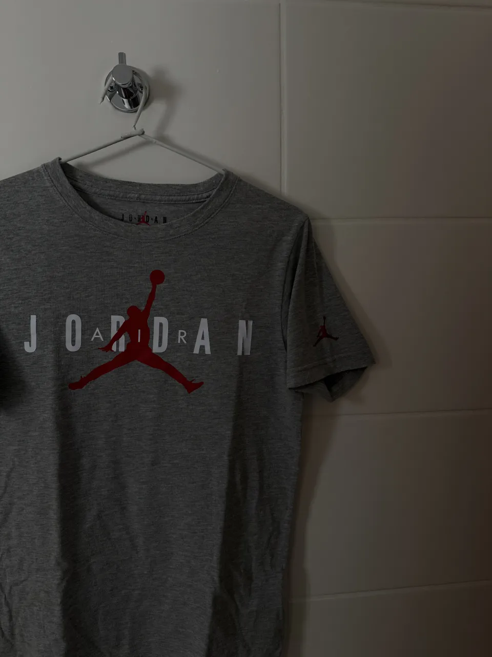 Remera gris de manga corta con estampa frontal de "JORDAN" en rojo y blanco. Logo de Jordan en rojo en la manga izquierda y en la parte superior del cuello.