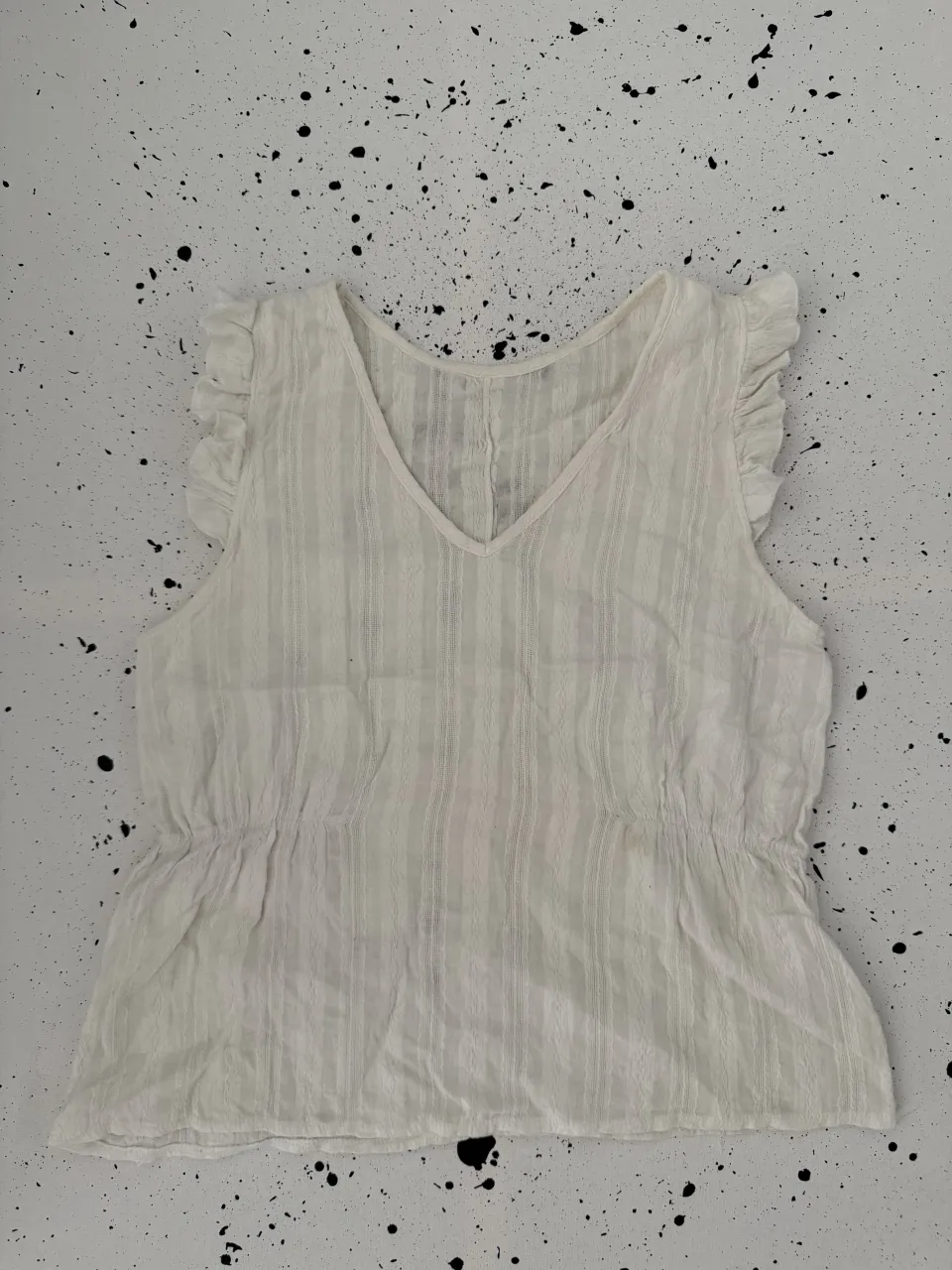 Remera blanca sin mangas con escote en V y detalle de fruncido en la cintura. Posee volados en los hombros. La tela tiene un tramado de finas rayas verticales.