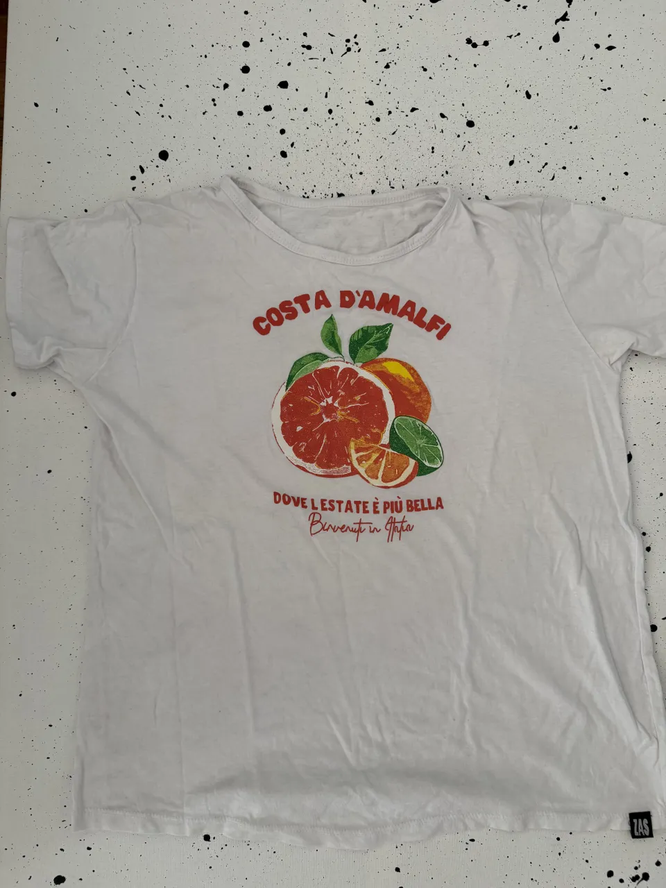 Remera blanca con estampa "COSTA D'AMALFI" en la parte delantera. La estampa incluye una naranja cortada, hojas verdes y texto en italiano. La estampa es de estilo vintage.