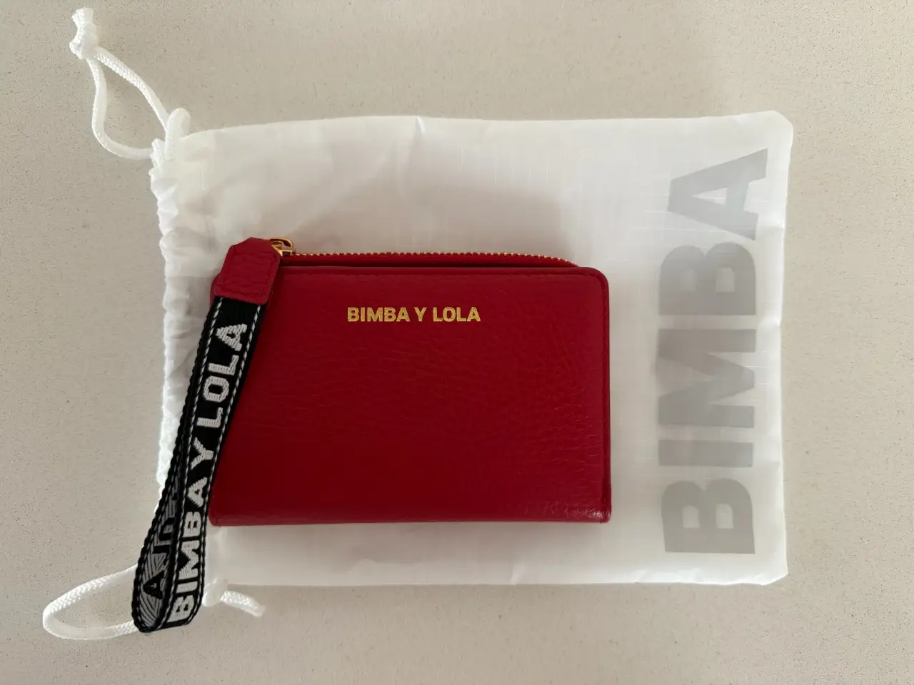 Billetera roja de cuero con cierre y correa de mano con la marca BIMBA Y LOLA. Incluye bolsa de tela blanca con el logo de la marca.