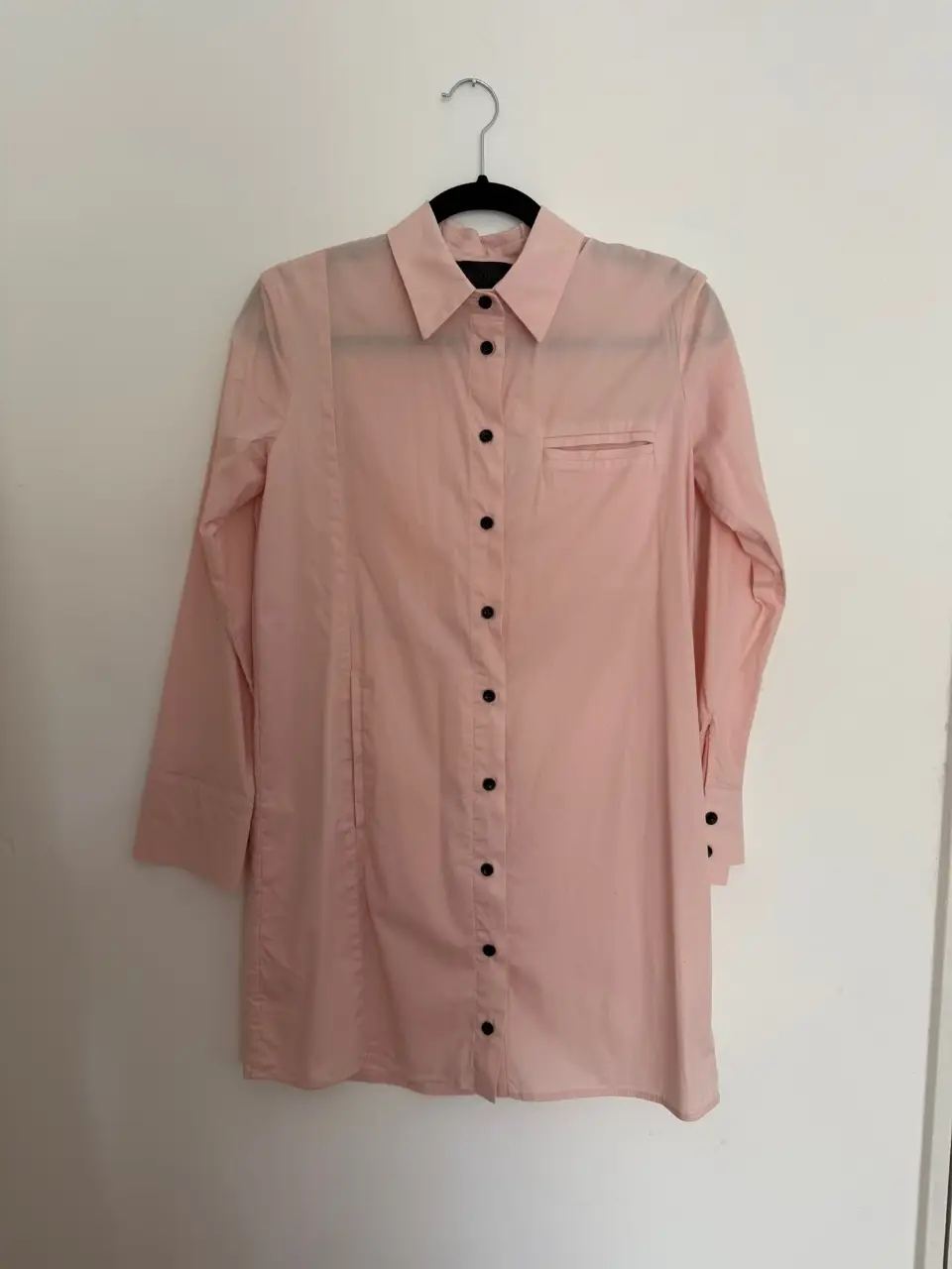 Camisa larga de color rosa con botones negros. Tiene un bolsillo en el pecho y mangas largas con puños.