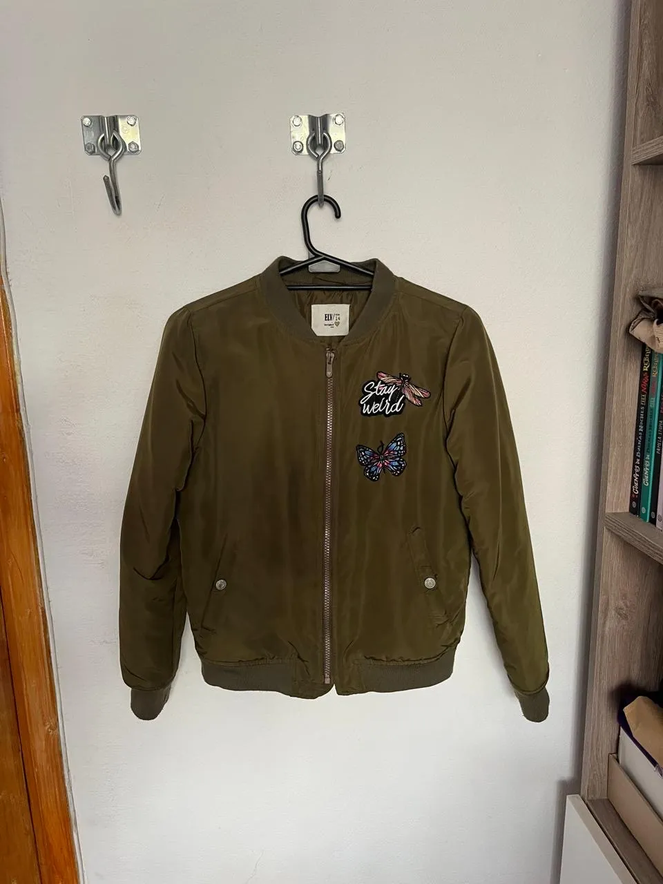 Campera bomber color verde militar. Tiene un cierre al frente y dos bolsillos con botones a los costados. En el frente tiene un aplique de mariposa y otro de una abeja con la frase "Stay weird". En la espalda tiene la frase "Take it easy" estampada en blanco. Marca ELV