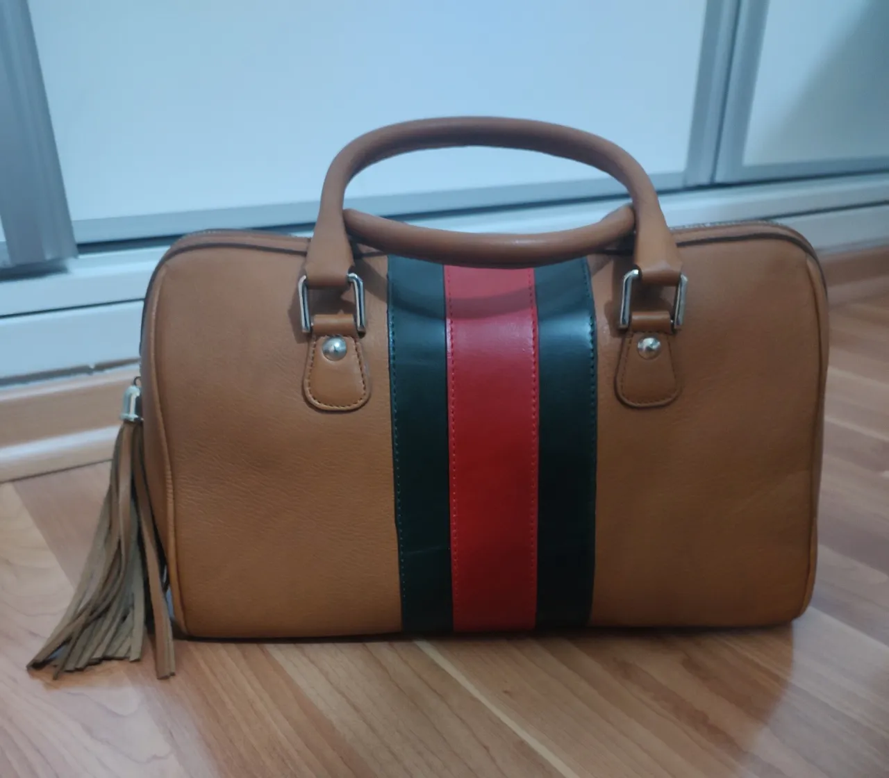 Bolso de mano para mujer, color marrón con franja central verde, roja y negra. Detalles de diseño con flecos y asas superiores. Ideal para un look casual y elegante.