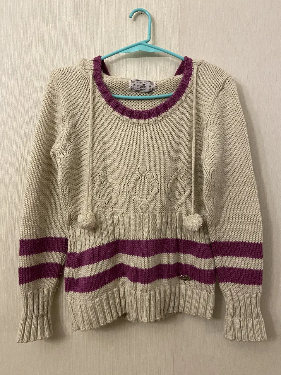 Sweater de mujer tejido en color crudo con detalles en violeta. Diseño con capucha y pompones, ideal para un look casual y abrigado.