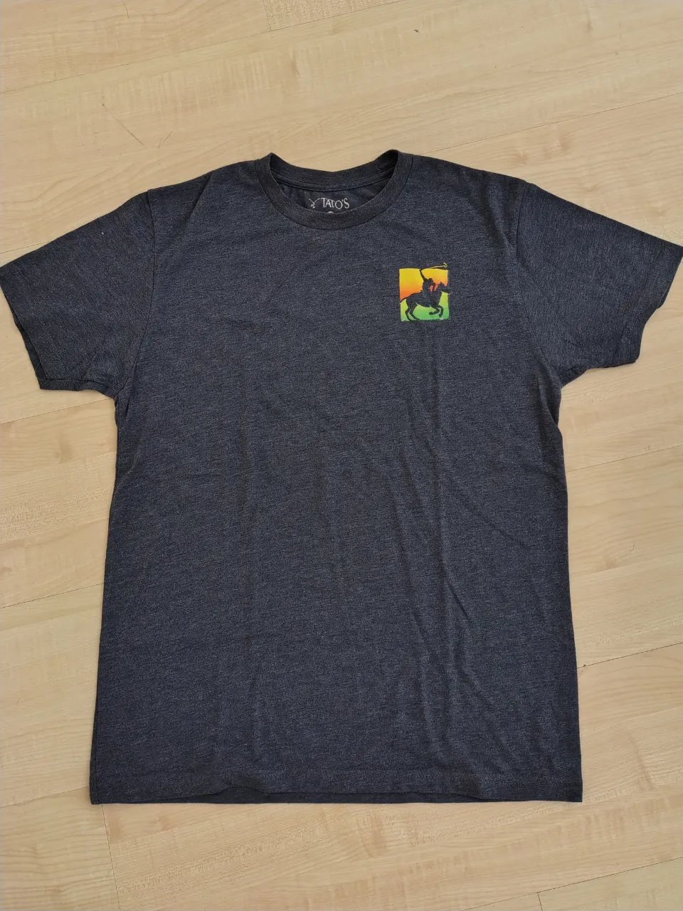Remera de manga corta de color gris oscuro. Al frente, tiene un pequeño estampado de un logo con un jinete y un caballo. En la espalda, el estampado es más grande y dice "SUNSET POLO at Great Meadow".