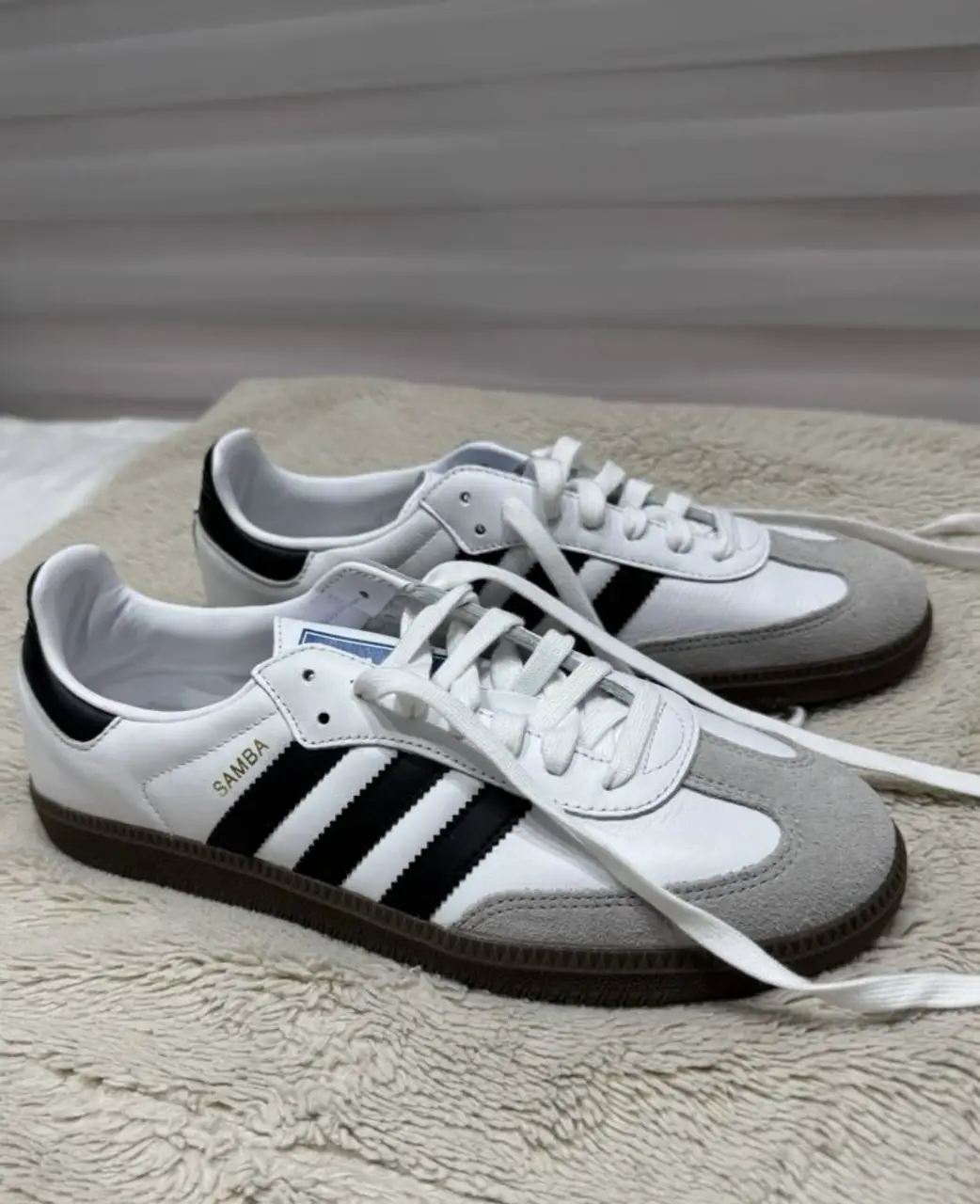 Zapatillas Adidas Samba blancas con detalles en negro y gris. Tienen las tres tiras características de la marca y suela de goma marrón. Un clásico atemporal para un look casual y urbano.