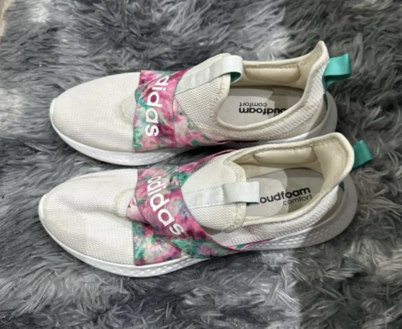Zapatillas Adidas blancas con detalles florales en la banda central. Diseño slip-on con suela blanca y detalles en verde turquesa. Ideales para un look casual y cómodo.