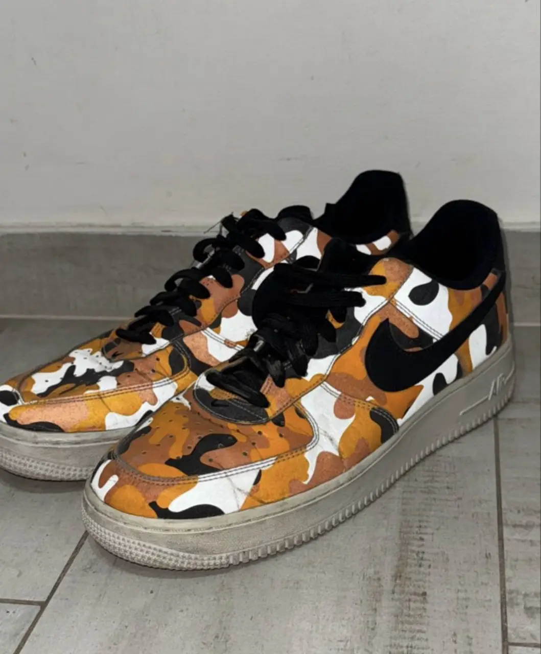 Zapatillas Nike Air Force con estampado camuflado en tonos naranja, blanco y negro. Tienen detalles en negro y suela blanca. Ideales para un look urbano y moderno.