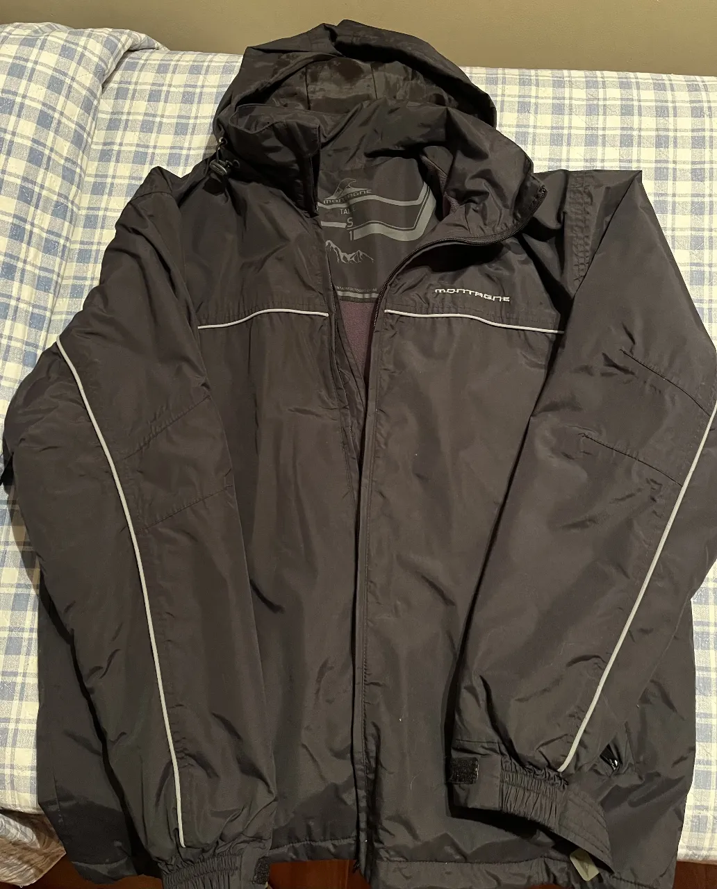 Campera Montagne negra con capucha, ideal para actividades al aire libre. Posee cierre frontal, bolsillos y detalles reflectantes. Perfecta para protegerte del viento y la lluvia.