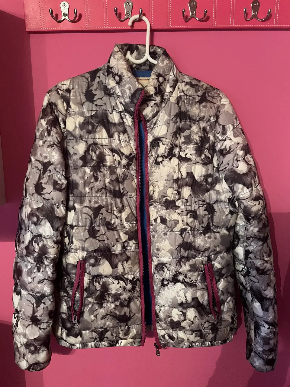 Campera acolchada con estampado floral en tonos grises y blancos, con detalles en rosa. Tiene cierre y bolsillos con cierre. Interior azul.
