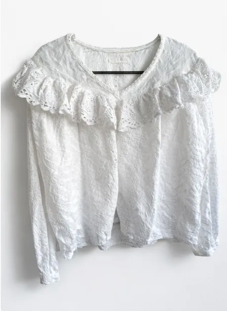 Blusa blanca con mangas largas y detalles de volados en el cuello y los hombros. Posee un tejido texturizado con un patrón floral o de hojas.