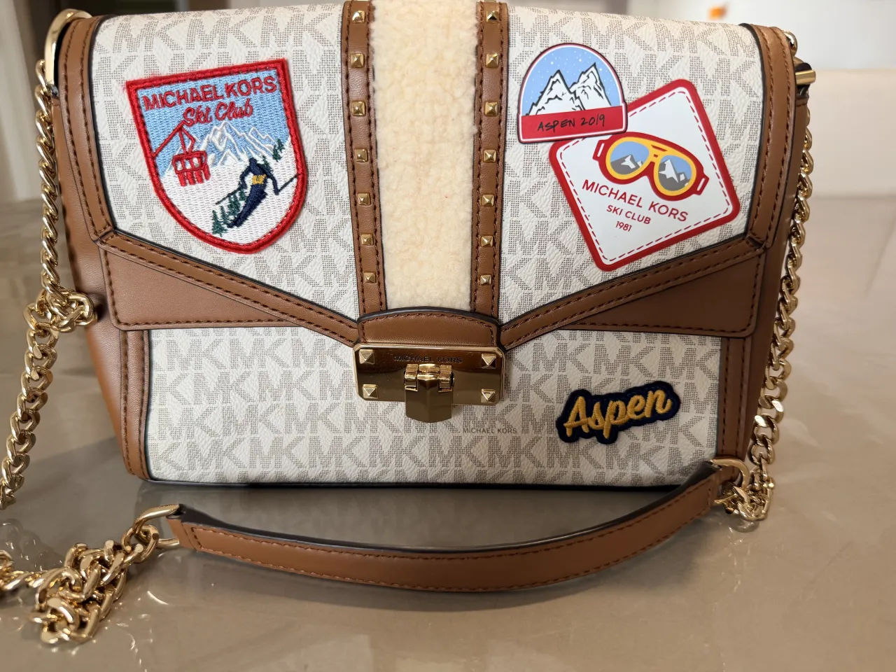 Bolso Michael Kors con diseño de parches de ski y logo MK. Cadena dorada y detalles en cuero marrón. Ideal para un look casual y divertido.