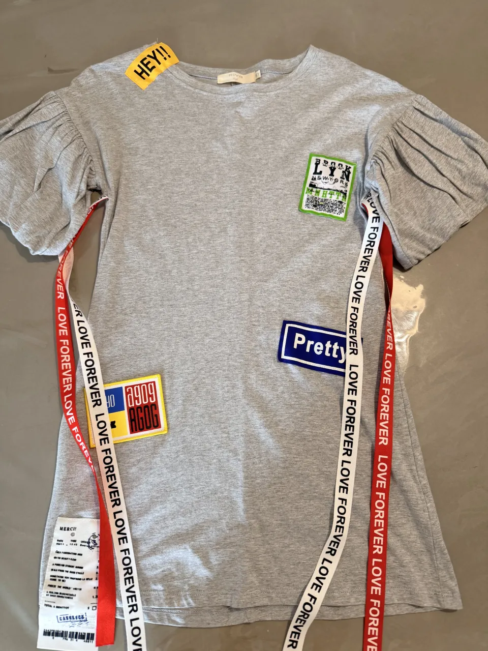 Remera gris con mangas abullonadas y apliques divertidos. Tiene cintas con la inscripción "LOVE FOREVER" a los costados. Ideal para un look casual y original.