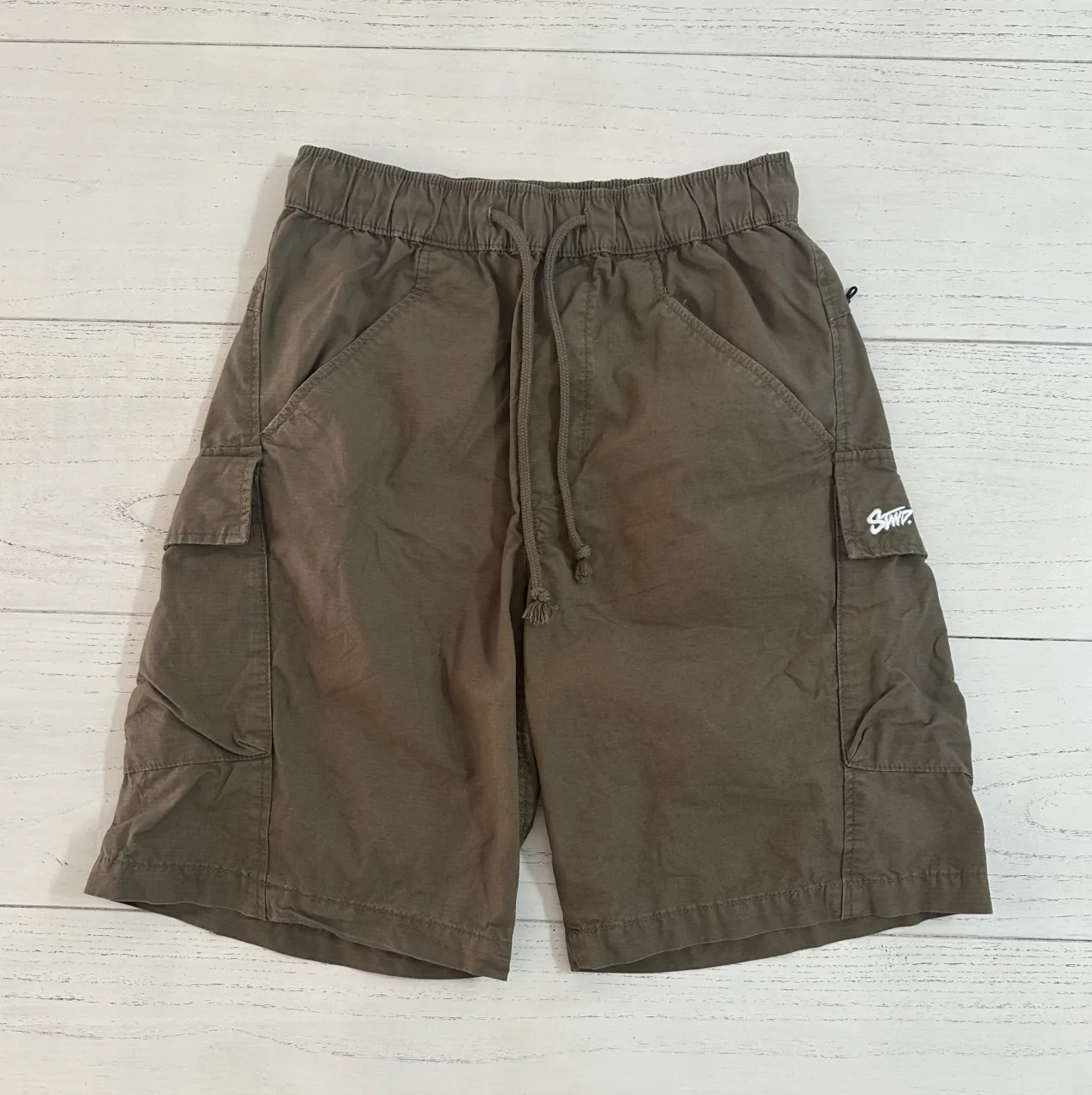 Shorts cargo de color marrón con cordón ajustable en la cintura y bolsillos laterales con cierre. Presenta un logo blanco estampado en el bolsillo lateral derecho.