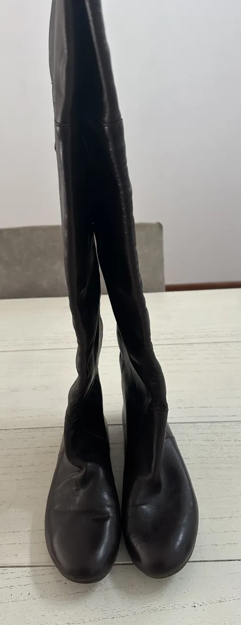 Botas de cuero marrones, caña alta, con cierre lateral y detalle de moneda colgante.