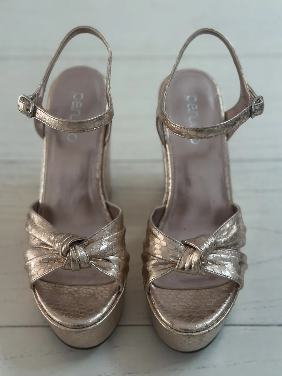 Sandalias doradas con plataforma y taco cuña. Tienen tiras finas con hebilla en el tobillo y una tira delantera con nudo.