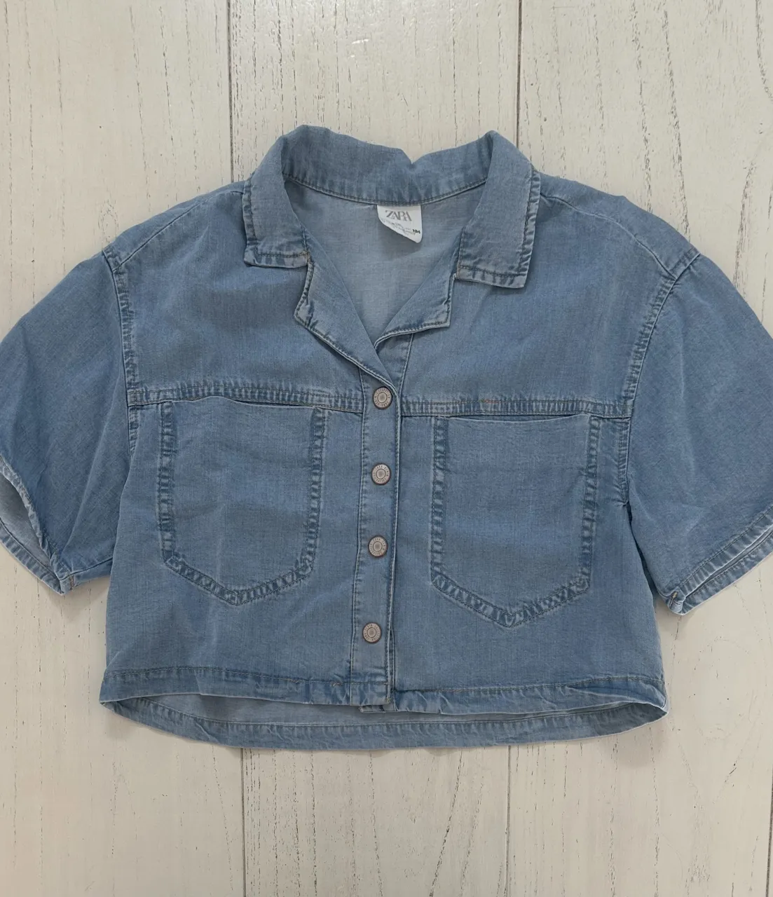 Camisa de jean manga corta con cuello y botones al frente. Posee dos bolsillos con tapa en el frente.