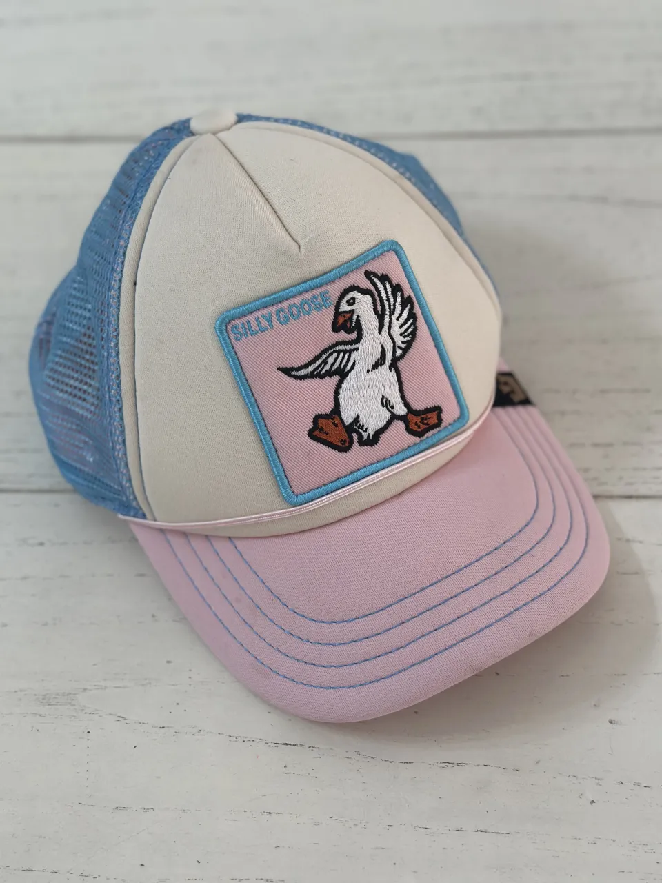 Gorra trucker con parche bordado de un ganso con la inscripción "Silly Goose". La visera es de color rosa con costuras y la parte trasera es de red azul. La parte frontal es de color blanco roto.