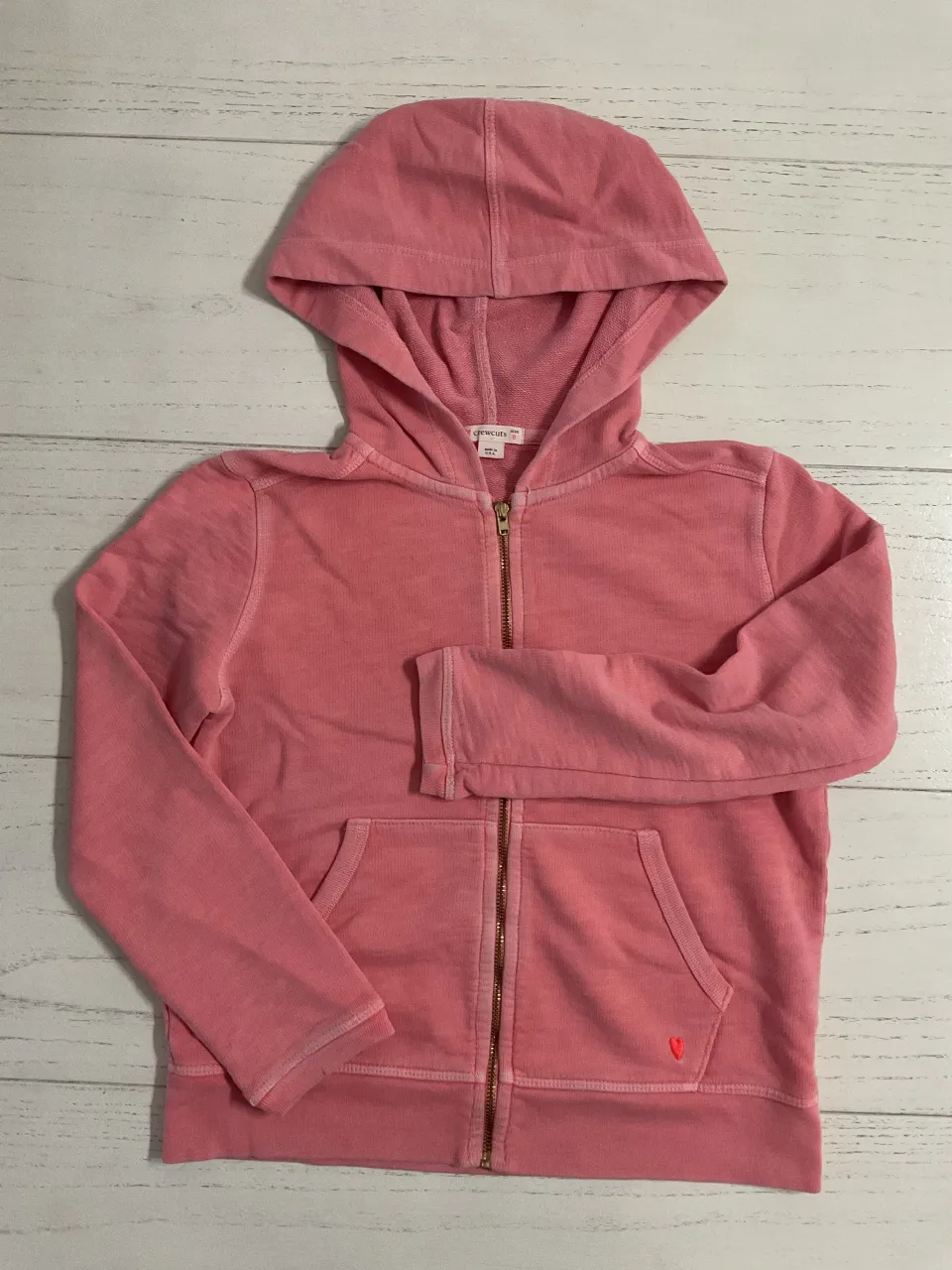 Buzo con capucha y cierre completo. Tiene bolsillos frontales tipo canguro y un pequeño corazón bordado en la parte inferior izquierda.