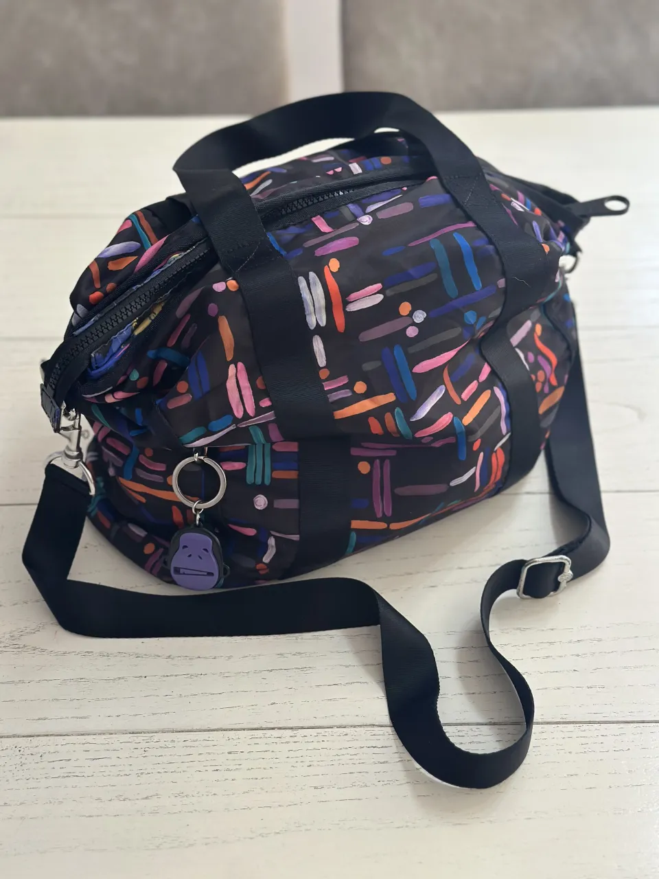 Bolso de mano y/o al hombro con correa ajustable. Posee múltiples compartimentos internos y externos con cierre. Estampado multicolor sobre fondo negro.