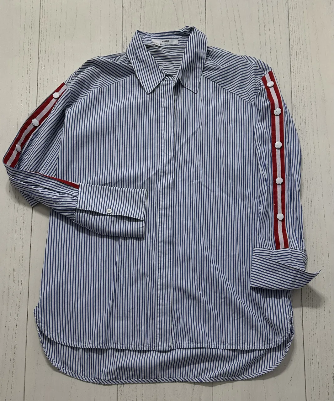 Camisa a rayas verticales azules y blancas. Posee detalles de tiras rojas con botones blancos en las mangas. Cuello clásico.