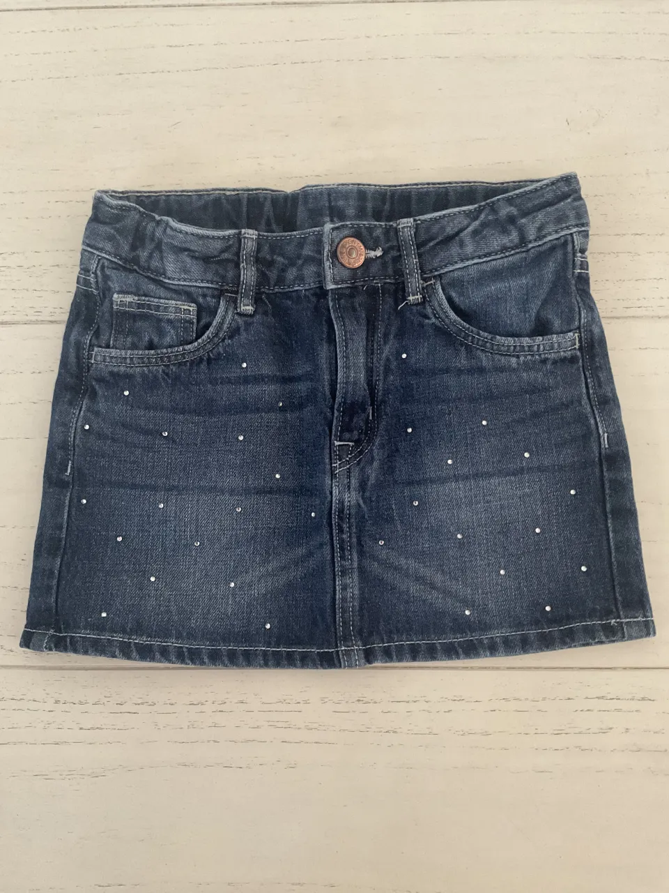 Pollera de jean azul con detalles de strass plateados. Tiene bolsillos delanteros y traseros.