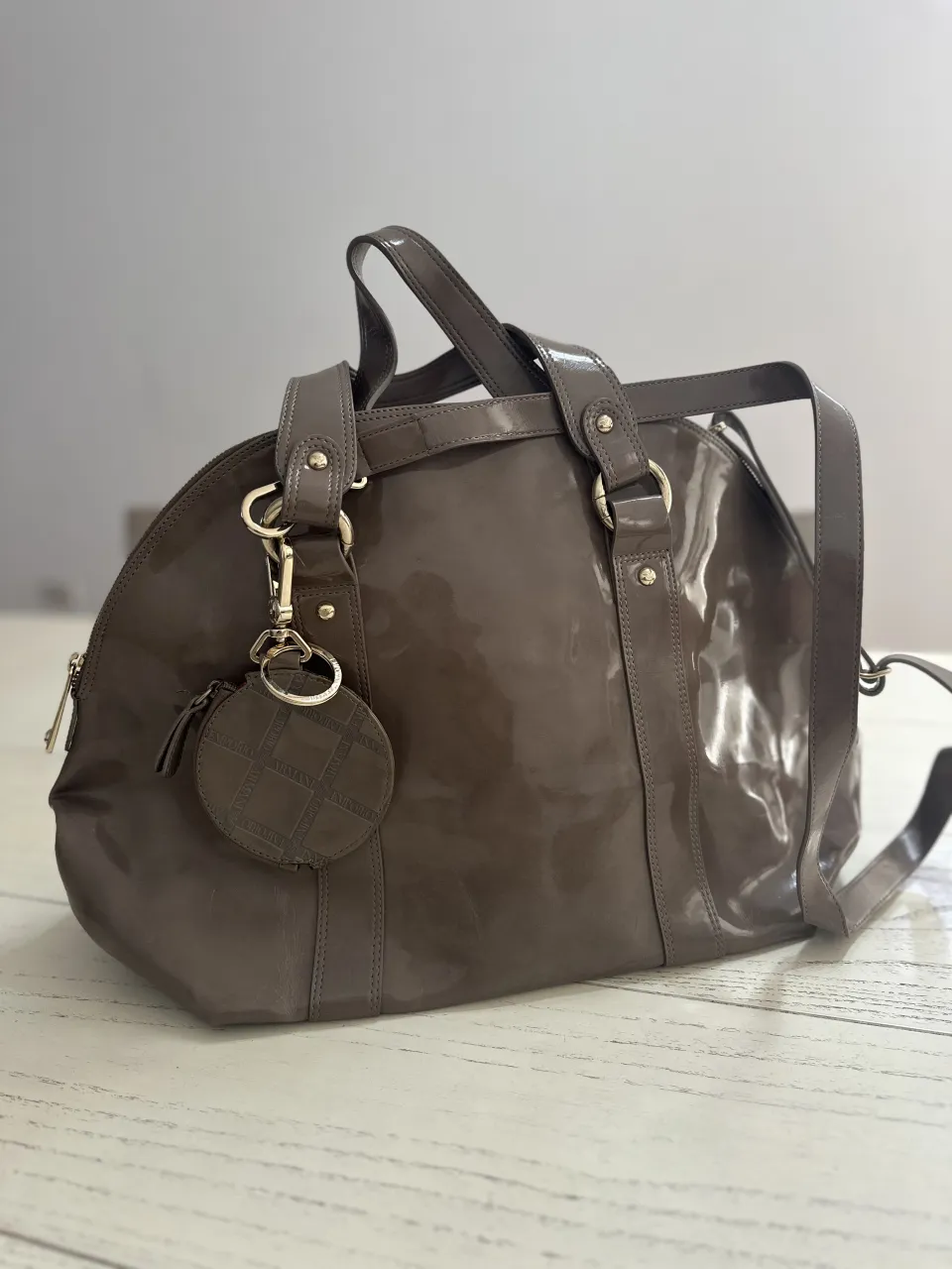 Bolso de cuero sintético color marrón. Tiene dos asas cortas y una correa larga desmontable. Incluye un pequeño monedero colgante con un diseño a cuadros. El interior es de color rosa con un estampado de la marca.