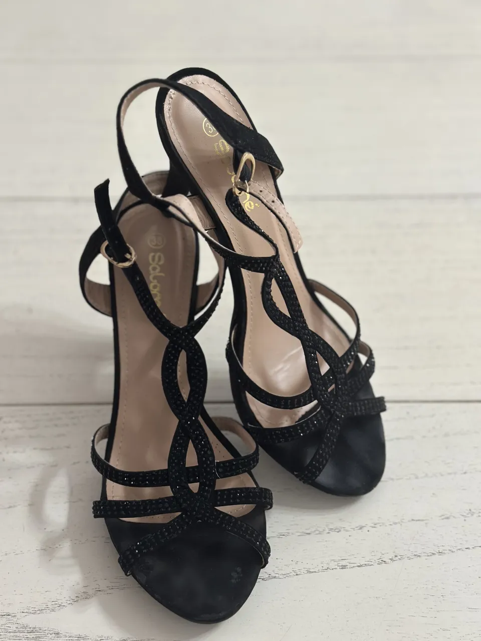 Sandalias negras con tiras finas adornadas con pedrería. Cierre de hebilla en el tobillo. Taco alto.