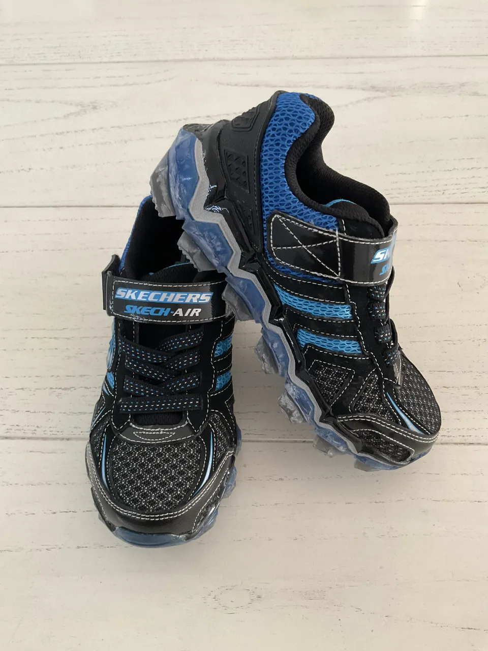 Zapatillas deportivas para niño, marca Skechers, con abrojo y cordones. Diseño en negro y azul con detalles reflectantes y suela con relieve.